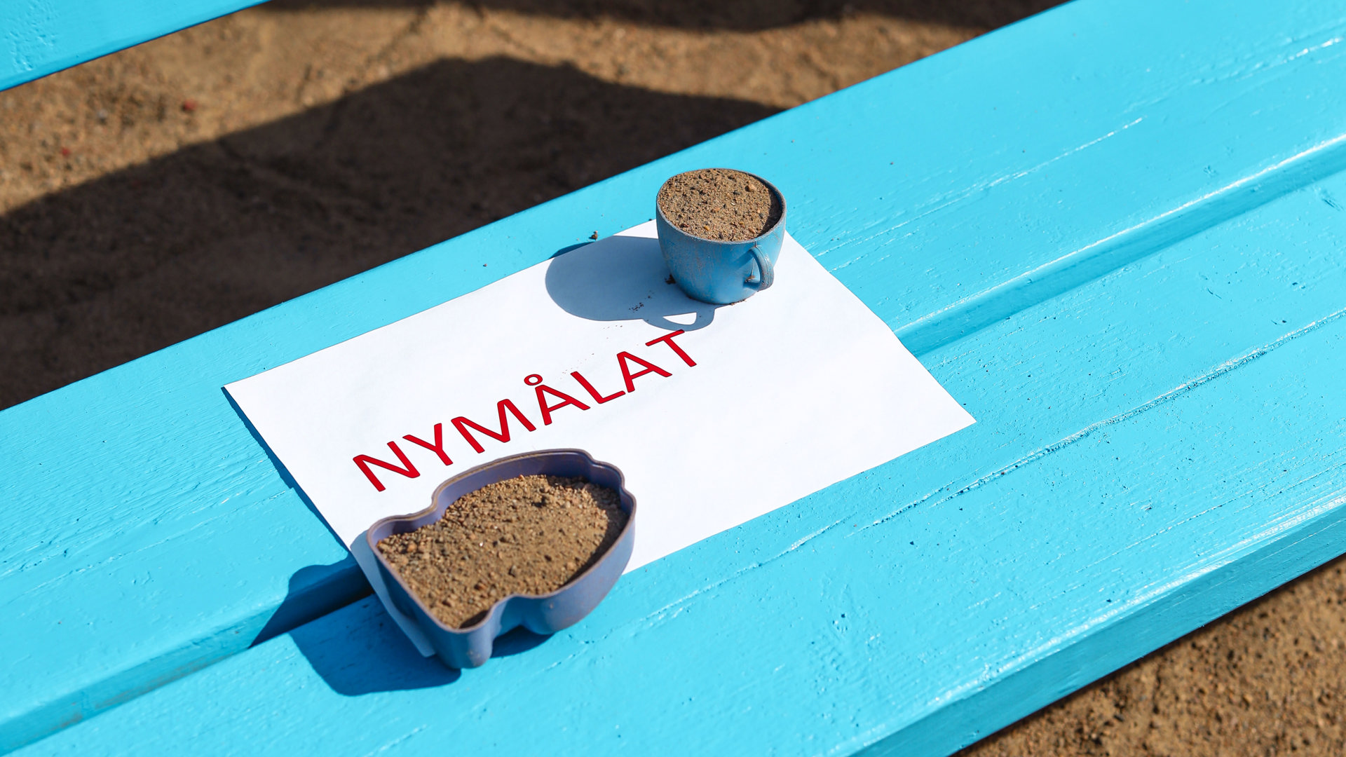 Blå träbänk med en lapp på där det står ´nymålat´med röda bokstäver. Under bänken syns sand. 