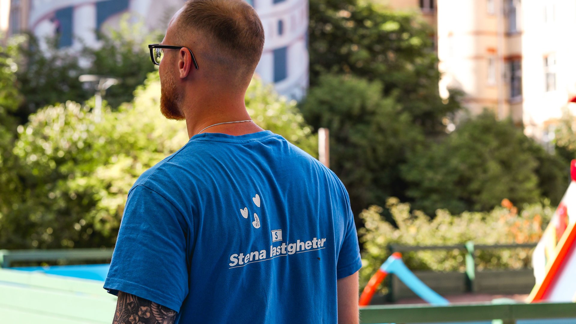En person sedd bakifrån, under ett parasoll en sommardag, iförd en blå t-shirt med texten 'Stena Fastigheter' på ryggen. I bakgrunden syns en lekplats som solen skiner på.