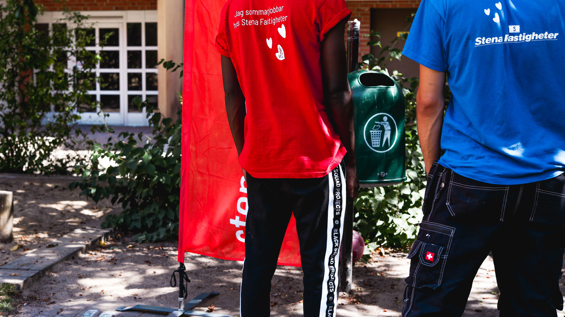 Två personer står vända från kameran. Den ena har en röd t-shirt på sig med texten ´Jag sommarjobbar hos Stena Fastigheter`. Den andra har en blå t-shirt med Stena Fastigheters logga på ryggen. De befinner sig utomhus en solig dag. 