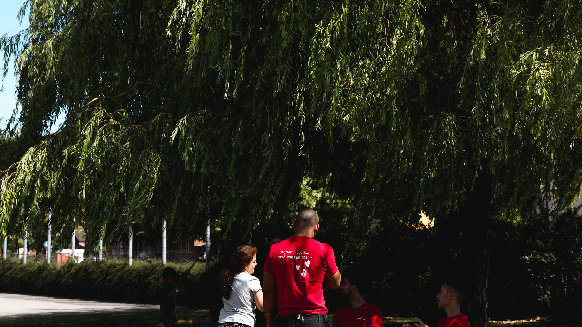 Fem personer befinner sig i skuggan under ett stort träd en solig sommardag. De verkar ha en konversation och en av personerna håller en läskburk i handen. Tre av personerna bär röda t-shirts med budskapet att de sommarjobbar hos Stena Fastigheter.  I bakgrunden syns två barn leka under samma träd. 