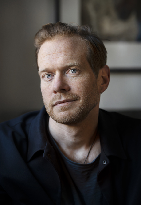 Närbild på Johan Bävman,fotograf. Han kollar in i kameran och har ett litet snett leende med stängd mun. 