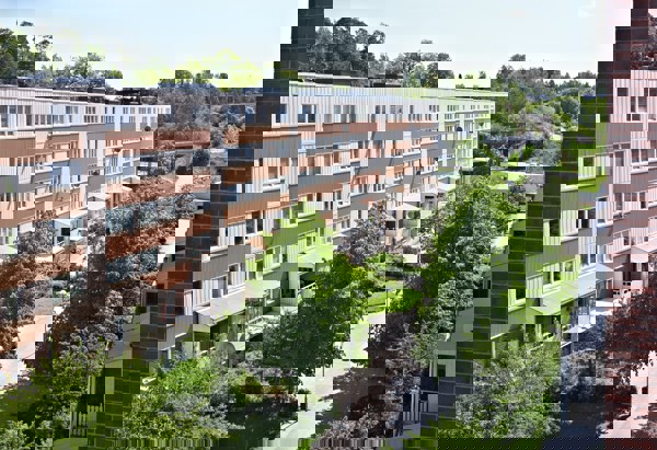 Lägenhetshus i Fisksätra, Stockholm en solig dag. Bakom huset syns grönska och träd. Framför huset finns gräsplättar, fler träd och lyktstolpar. Bilden är tagen ovanifrån från ett hus mittemot 