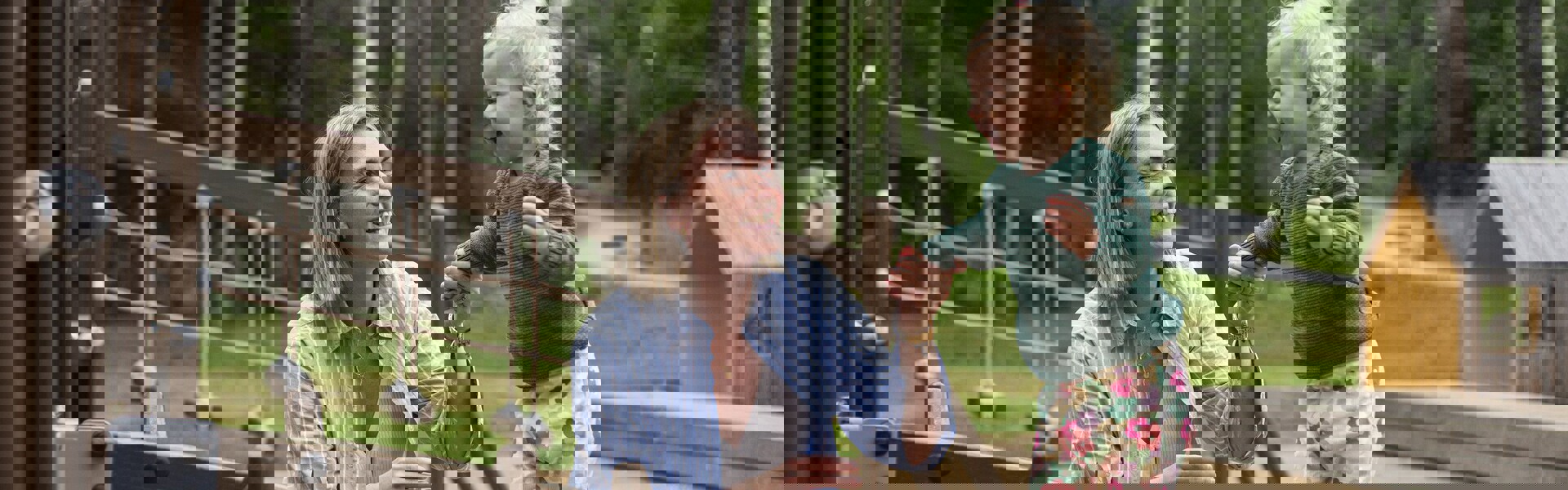 En leende vuxen håller ett litet barn i händerna när barnet balanserar på en lekställning på en lekplats utomhus, omgiven av träd.