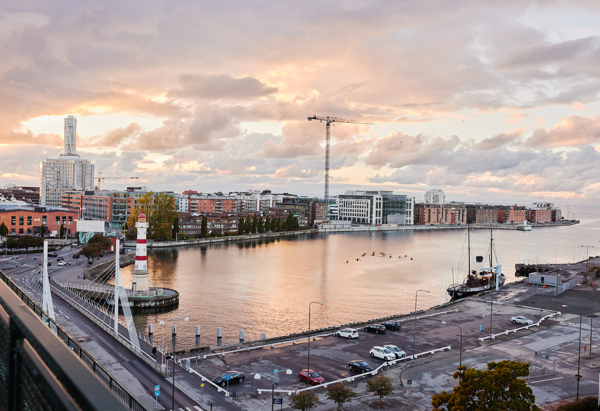 Bild tagen en sommarkväll när solen är på väg ner. Himlen är rosa med fina moln. I mitten av bilden syns vatten, och på ena sidan finns hus och byggnader. En bro sträcker sig över vattnet till andra sidan, där det finns en parkering med några bilar och små träd