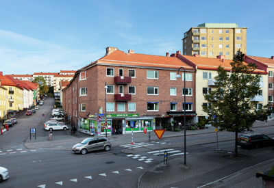 Stadsbild av en väg där två bilar kör. På andra sidan vägen syns röda och gula lägenhetshus med röda tak. På hörnet av ett av husen finns en affär med grön skyltning.