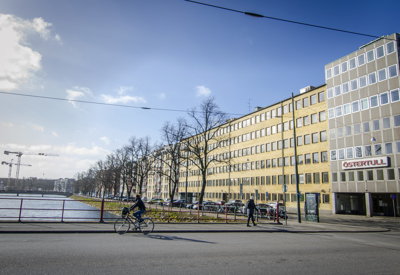 En stadsvy med en bred gata i förgrunden där en cyklist och en fotgängare rör sig. Till höger syns en gul och vit kontors- eller bostadsbyggnad med skylten "ÖSTERTULL" på fasaden. Till vänster om byggnaden sträcker sig en rad med träd längs ett vattendrag, och flera bilar står parkerade. I bakgrunden syns en bro och två byggkranar. Himlen är blå med några moln, och solen skiner.