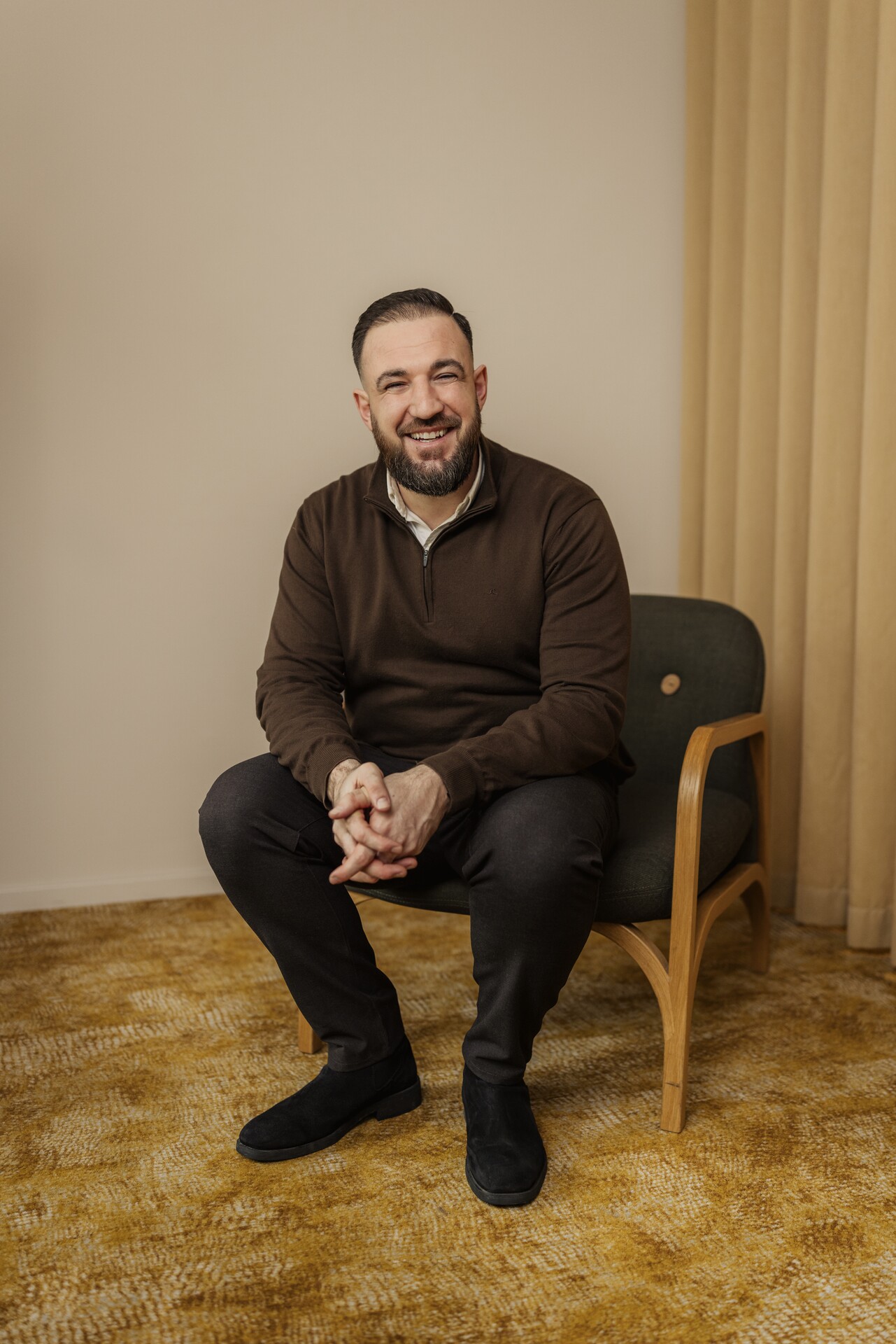 En leende man, Bledar, sitter avslappnat på en stol i ett varmt inrett rum och ser rakt in i kameran, vilket förmedlar trygghet; bilden illustrerar Stena Fastigheters arbete med social hållbarhet genom närvarande relationsförvaltning, där starka relationer till hyresgäster och lokalsamhälle är centrala för att skapa trivsamma och trygga bostadsområden