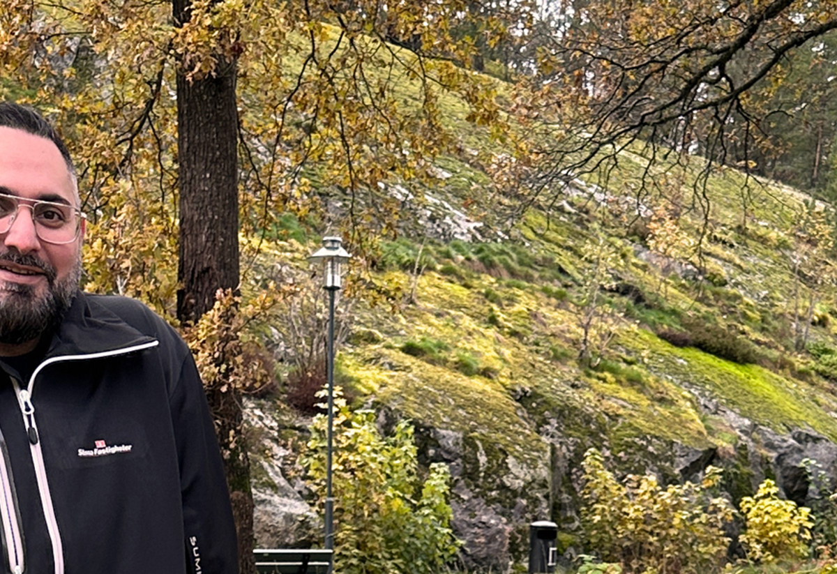En person med skägg och glasögon står utomhus i en skogsmiljö med höstfärger. Personen bär en mörk jacka, och bakgrunden visar träd och en mossbeklädd sluttning.