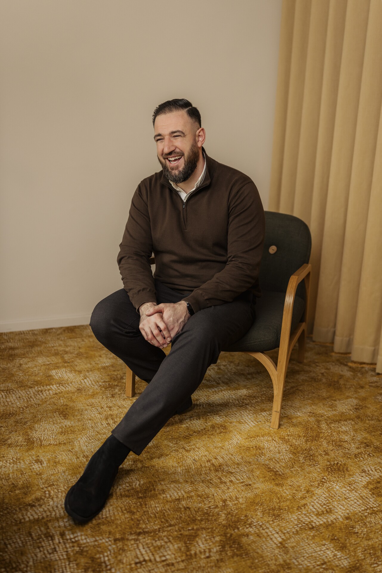  En person sitter avslappnat på en stol i ett varmt inrett rum och ler åt något utanför bild, vilket ger en känsla av trygghet; bilden anknyter till Stena Fastigheters arbete med social hållbarhet och relationsförvaltning, där närvaro och goda relationer i bostadsområden står i fokus.