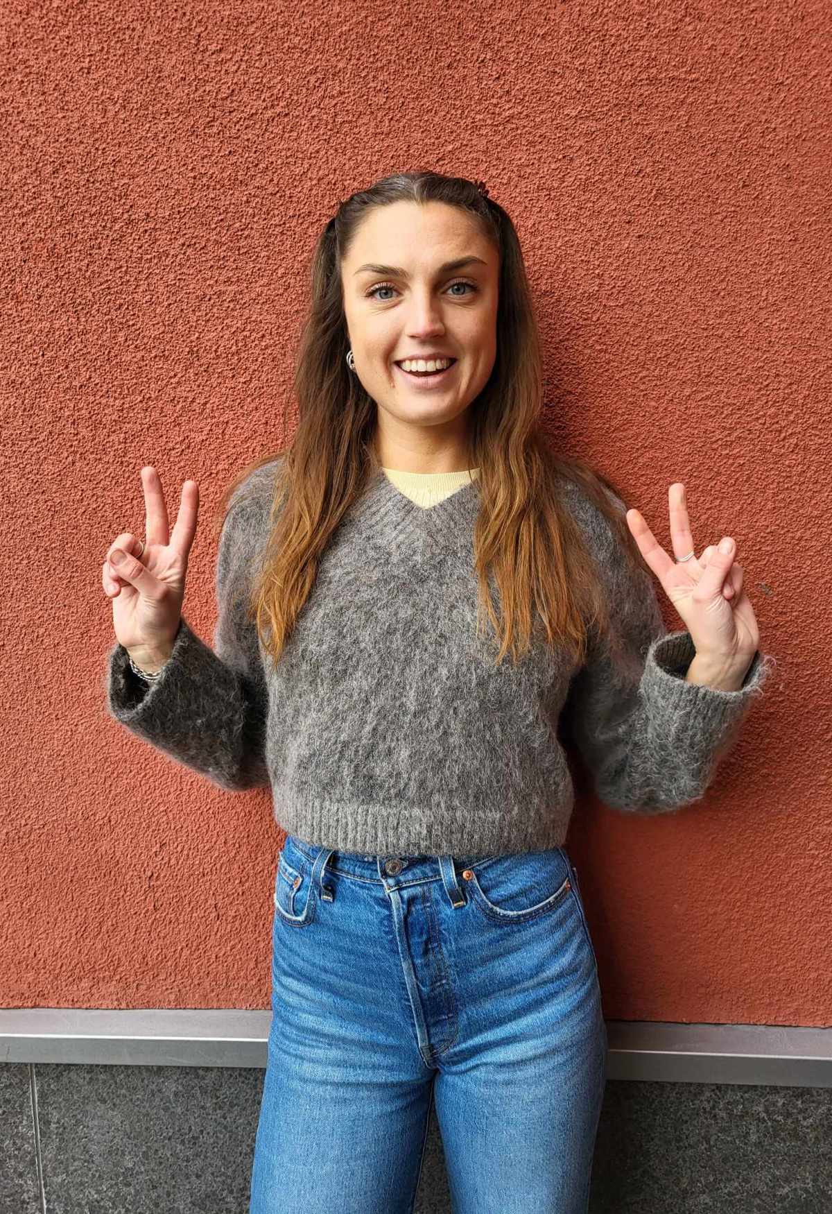 Glad ung tjej med långt brunt hår som gör peace-tecknet med fingrarna, tittar in i kameran och ser energisk ut.