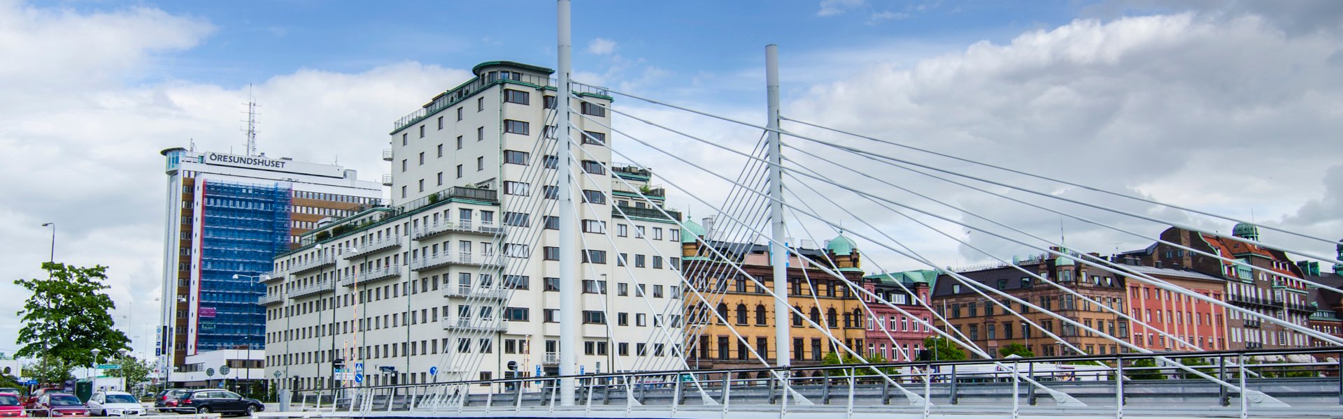 Vatten nedanför en bro, med en rad byggnader i olika färger ovanför. På hörnet ligger ett högt vitt hus som är Stena Fastigheters kontor i Malmö. Himlen ovanför husen är blå med moln.