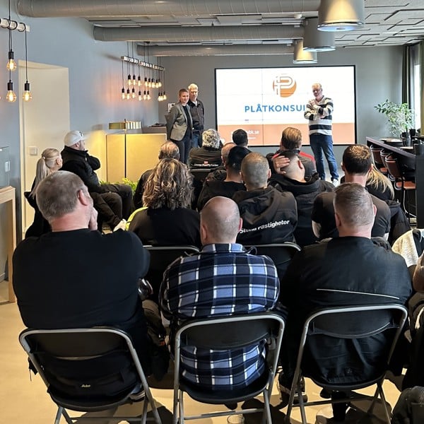 Fastighetsvärdar på Stena Fastigheter sitter på huvudkontoret för en träff och tittar på en presentationsskärm. Skärmen visar logotypen och namnet 'PLÅTKONSULT' tillsammans med ytterligare text och grafik. Tre personer står längst fram i rummet och leder presentationen.