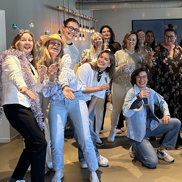 En grupp kollegor poserar tillsammans vid en festlig sammankomst. Personerna är klädda i festliga kläder, några bär glittriga plagg och accessoarer som glitter och huvudband