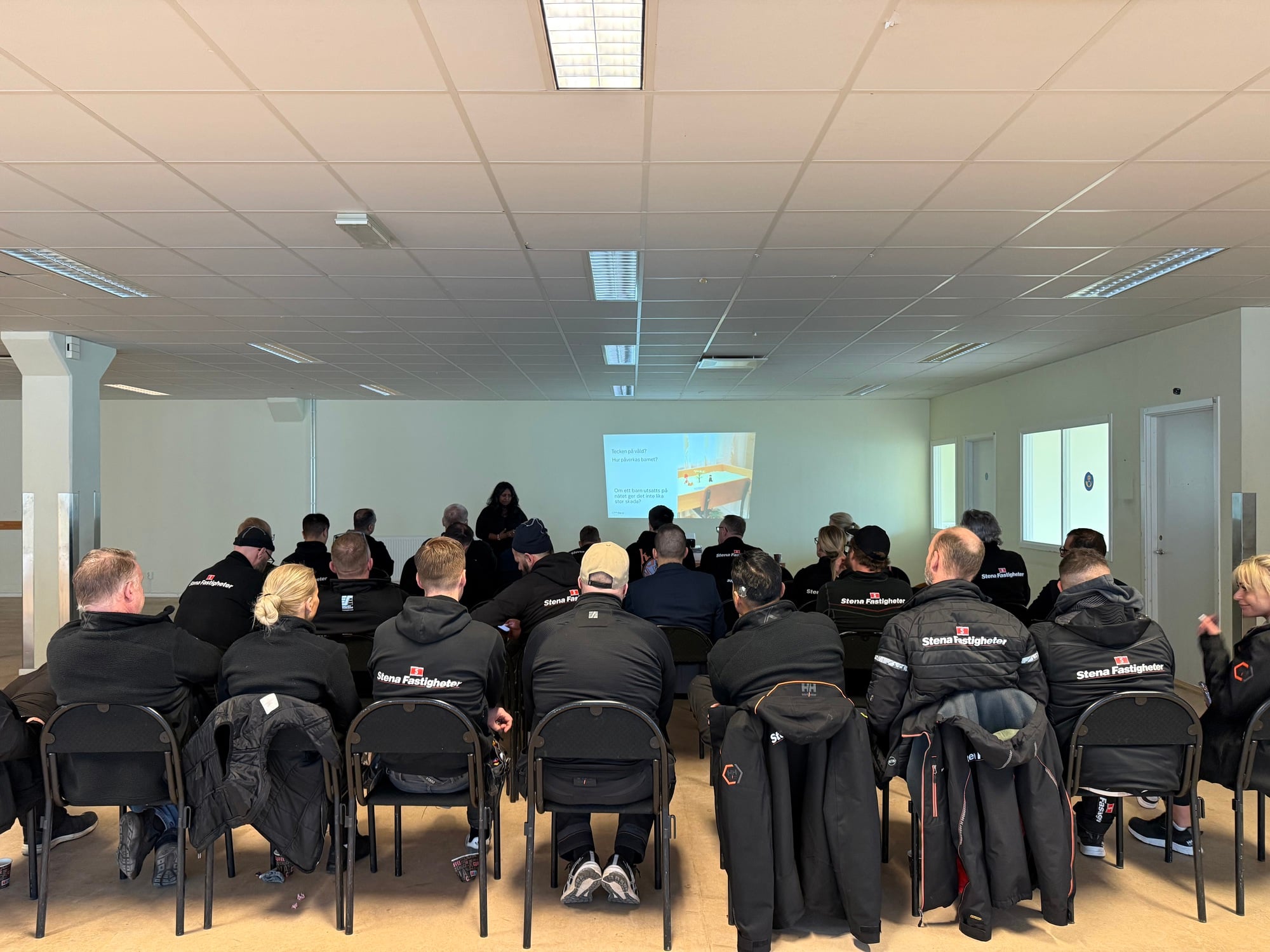 En grupp personer sitter i rader i ett konferensrum och lyssnar på en presentation längst fram i lokalen. Flera av deltagarna bär svarta jackor med Stena Fastigheters logotyp på ryggen.