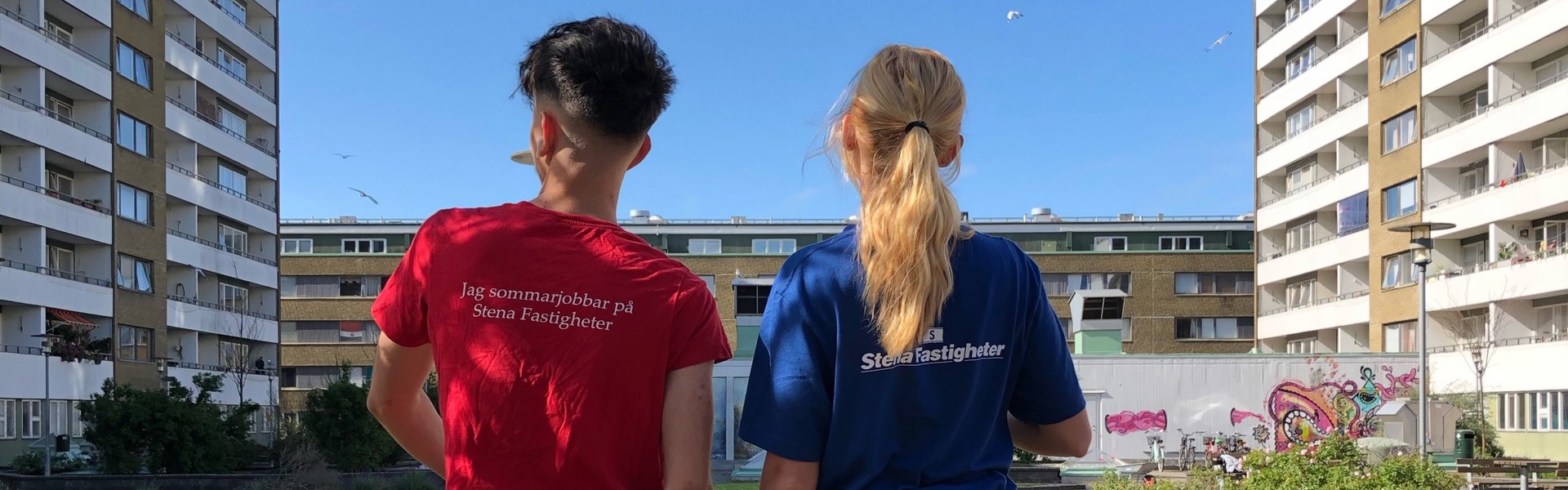 Två personer står med ryggen mot kameran och kollar ut över ett bostadsområde i Malmö. Vid sidorna och framför dem finns lägenhetshus. I mitten syns odlingsytor med gröna växter samt lila och rosa blommor. Den ena personen bär en röd t-shirt med texten 'Jag sommarjobbar hos Stena Fastigheter', och den andra har en blå t-shirt med Stena Fastigheters logga på ryggen