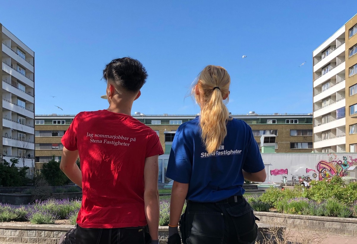 Två personer står med ryggen mot kameran och kollar ut över ett bostadsområde i Malmö. Vid sidorna och framför dem finns lägenhetshus. I mitten syns odlingsytor med gröna växter samt lila och rosa blommor. Den ena personen bär en röd t-shirt med texten 'Jag sommarjobbar hos Stena Fastigheter', och den andra har en blå t-shirt med Stena Fastigheters logga på ryggen
