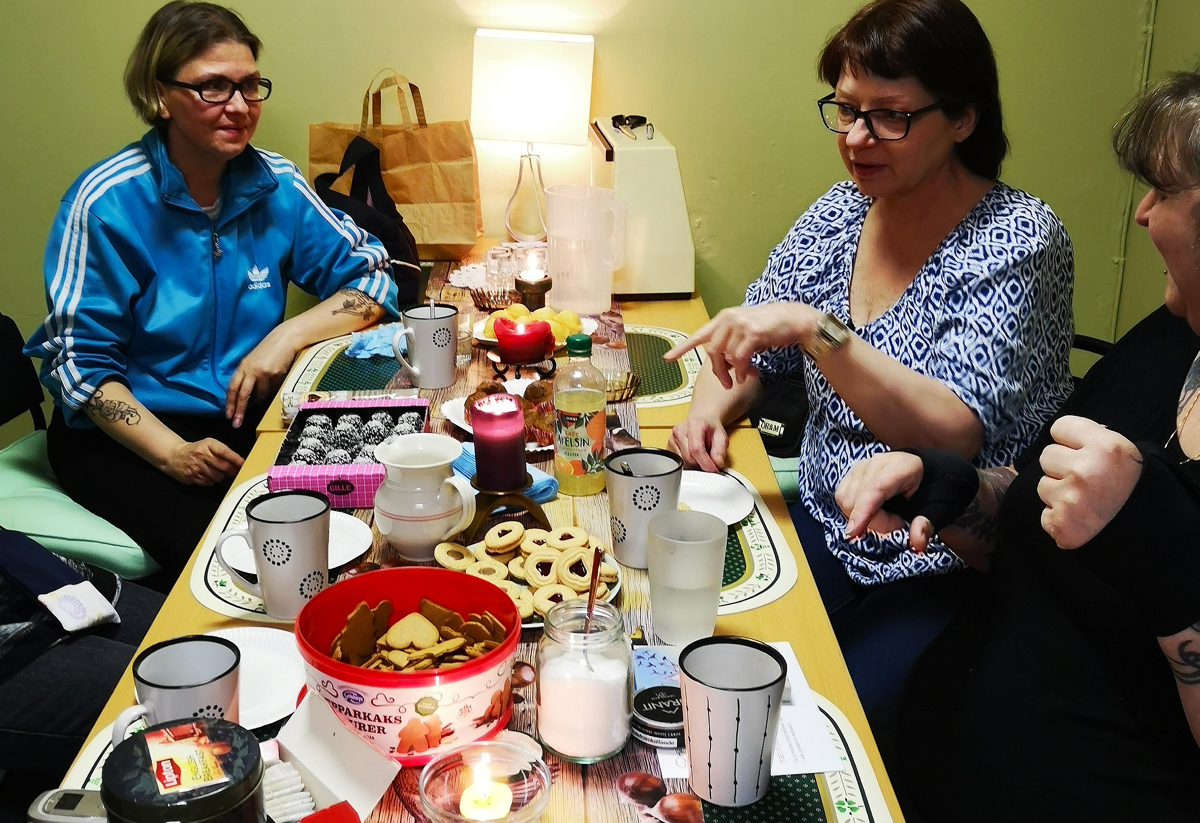 Tre personer sitter runt ett bord i en mysig miljö. Bordet är dukat med kakor, chokladbollar och drycker. En tänd lampa och ett levande ljus skapar en varm stämning. Personerna verkar vara engagerade i ett samtal och njuter av fikat tillsammans.