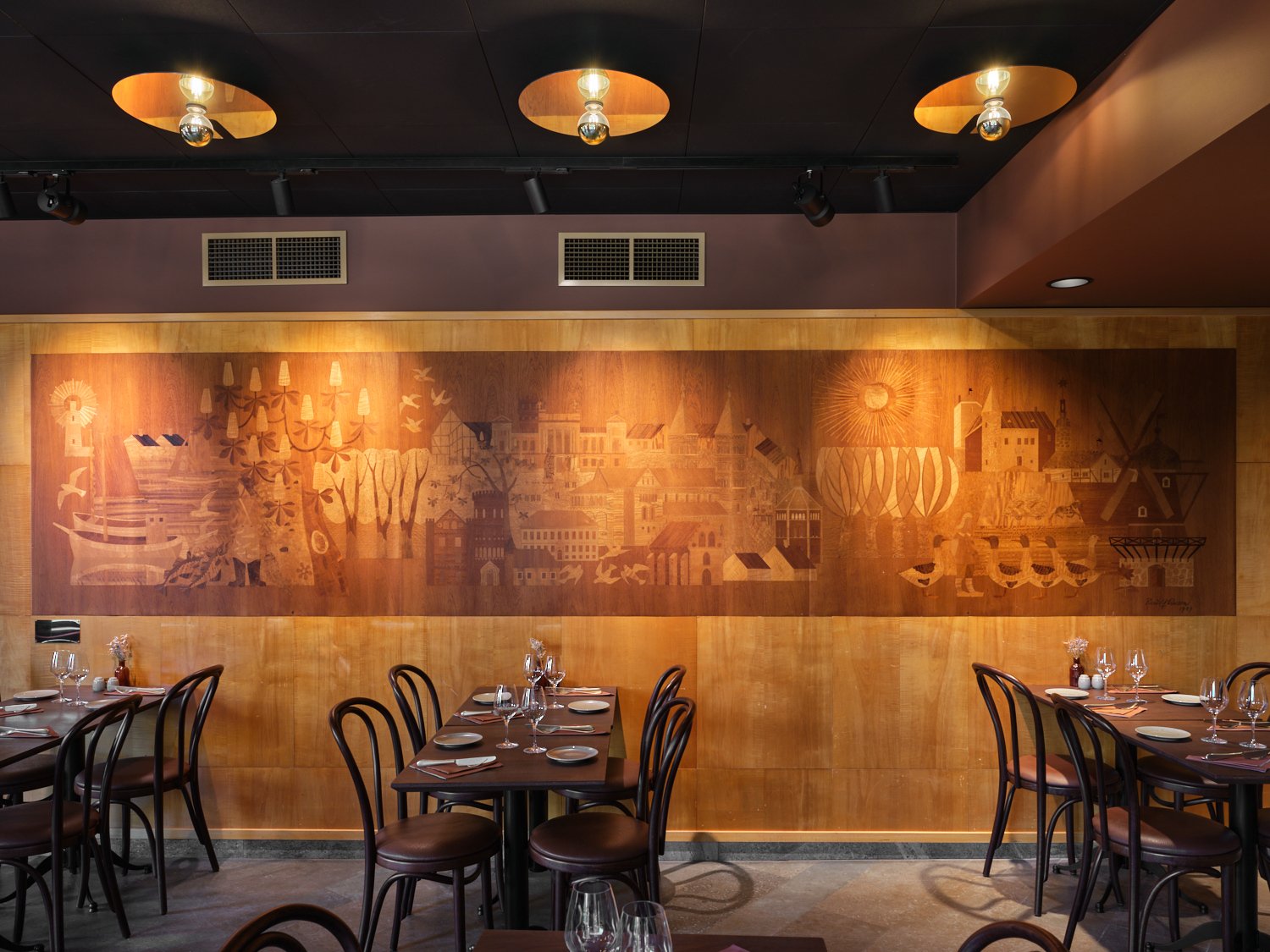 En varmrestaurangmilj&ouml; med m&ouml;rka tr&auml;stolar och dukade bord l&auml;ngs v&auml;ggarna. L&auml;ngs den bakre v&auml;ggen l&ouml;per ett stort, brunt konstverk i tr&auml; med inristade motiv av byggnader, tr&auml;d och figurer, upplyst av spotlights fr&aring;n det m&ouml;rka taket ovanf&ouml;r.