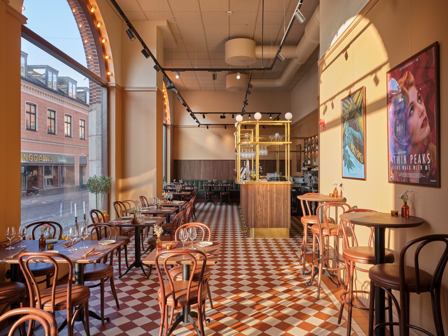 Ett ljust restaurangrum med rutigt r&ouml;dvitt klinkergolv, h&ouml;ga f&ouml;nster mot gatan och v&auml;ggar i varma gula toner. L&auml;ngs golvet st&aring;r sm&aring; dukade bord med b&ouml;jtr&auml;stolar, och l&auml;ngst in syns en bar med tr&auml;- och m&auml;ssingsdetaljer, medan f&auml;rgglada affischer pryder v&auml;ggen till h&ouml;ger.