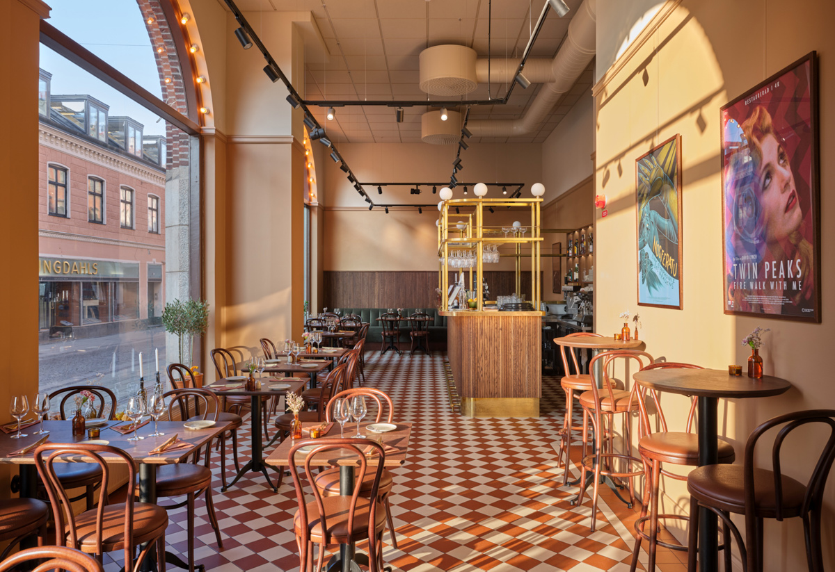 Ett ljust restaurangrum med rutigt rödvitt klinkergolv, höga fönster mot gatan och väggar i varma gula toner. Längs golvet står små dukade bord med böjträstolar, och längst in syns en bar med trä- och mässingsdetaljer, medan färgglada affischer pryder väggen till höger.
