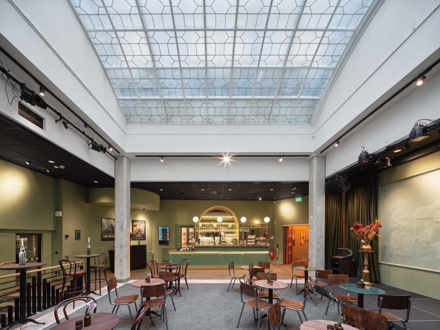 En rymlig foaj&eacute; med h&ouml;gt glastak som sl&auml;pper in ljus &ouml;ver ett &ouml;ppet s&auml;llskapsutrymme med sm&aring; runda bord och blandade tr&auml;stolar. L&auml;ngst bort syns en bar med gr&ouml;n front och v&auml;l upplyst serveringsdisk, omgiven av gr&ouml;na v&auml;ggar och scenutrustning, medan en h&ouml;g vas med blommor st&aring;r p&aring; ett sidobord till h&ouml;ger.