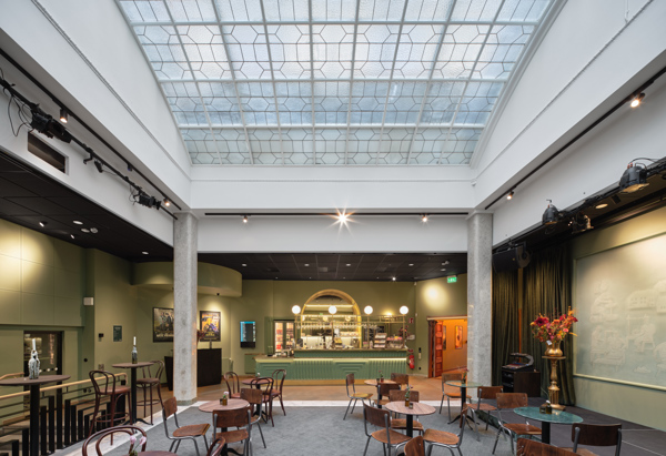 En rymlig foajé med högt glastak som släpper in ljus över ett öppet sällskapsutrymme med små runda bord och blandade trästolar. Längst bort syns en bar med grön front och väl upplyst serveringsdisk, omgiven av gröna väggar och scenutrustning, medan en hög vas med blommor står på ett sidobord till höger.