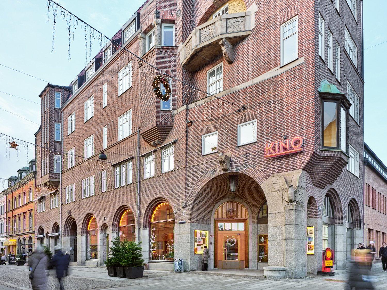 En exteri&ouml;rbild av en tegelbyggnad i h&ouml;rnl&auml;ge med stora rundb&aring;giga entr&eacute;er i gatuniv&aring; och flera v&aring;ningar ovanf&ouml;r. P&aring; fasaden sitter en neonskylt med texten &rdquo;KINO&rdquo;, och framf&ouml;r huset r&ouml;r sig m&auml;nniskor l&auml;ngs trottoaren, medan julbelysning och en gran i kruka skapar st&auml;mning p&aring; gatan.