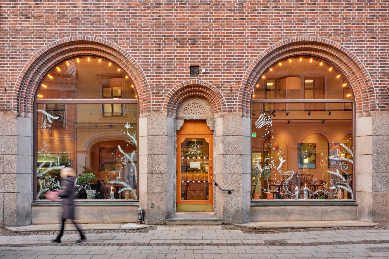 Exteri&ouml;rbild av en tegelbyggnad med tv&aring; stora rundb&aring;giga skyltf&ouml;nster och en tr&auml;d&ouml;rr i mitten, inramade av stenpelare. Genom f&ouml;nstren syns den varmt upplysta restaurang- eller caf&eacute;milj&ouml;n med bord och stolar, och p&aring; trottoaren utanf&ouml;r g&aring;r en f&ouml;rbipasserande person i r&ouml;relse.