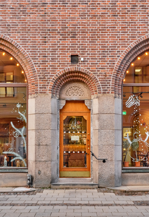 Exteriörbild av en tegelbyggnad med två stora rundbågiga skyltfönster och en trädörr i mitten, inramade av stenpelare. Genom fönstren syns den varmt upplysta restaurang- eller cafémiljön med bord och stolar, och på trottoaren utanför går en förbipasserande person i rörelse.