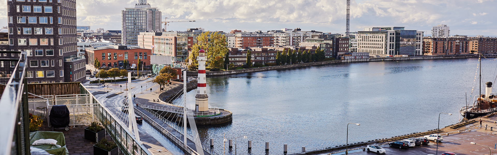En naturskön vy över Skeppsbron i Malmö. Bilden visar en hamn i centrum. En fyr är placerad nära vattenkanten. Flera byggnader, inklusive höghus och mindre byggnader, kantar vattnet. 