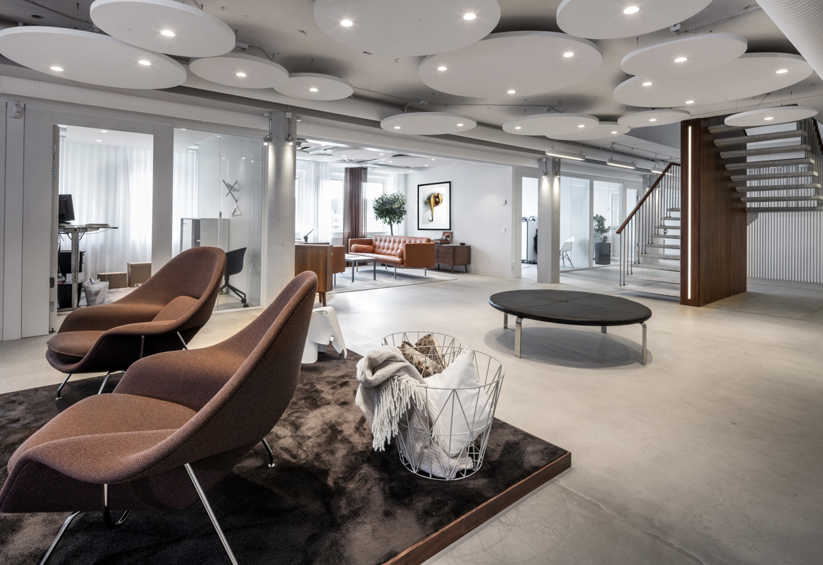 Modernt kontorsutrymme med stilren inredning. Rummet har bruna fåtöljer på en mörk matta, en rund sittmöbel och en trappa i bakgrunden. Taket är dekorerat med vita, cirkulära akustikplattor. Ljusa väggar och stora fönster ger ett luftigt och öppet intryck.