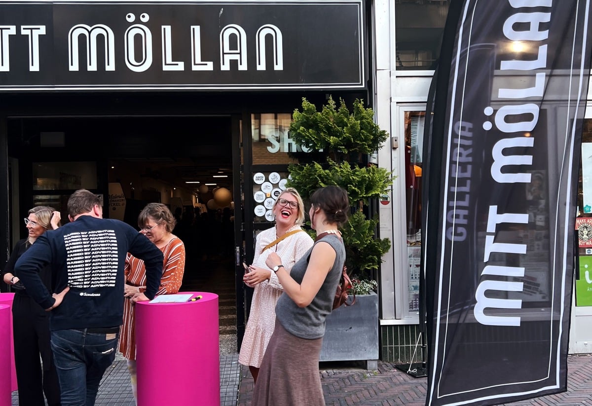Alt-text: "Folk står och samtalar utanför entrén till Mitt Möllan. Framför ingången finns rosa ståbord och en svart flagga med texten 'Gallerian Mitt Möllan'. På fasaden ovanför syns vita masker med olika ansiktsuttryck.