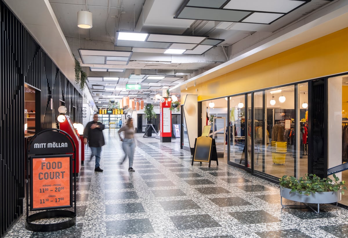 Inomhusbild av en modern galleria med människor som går förbi butiker och restauranger. Till vänster syns en skylt med texten 'Mitt Möllan Food Court'. Golvet har ett rutmönster och lokalen är ljus med stora fönster och taklampor