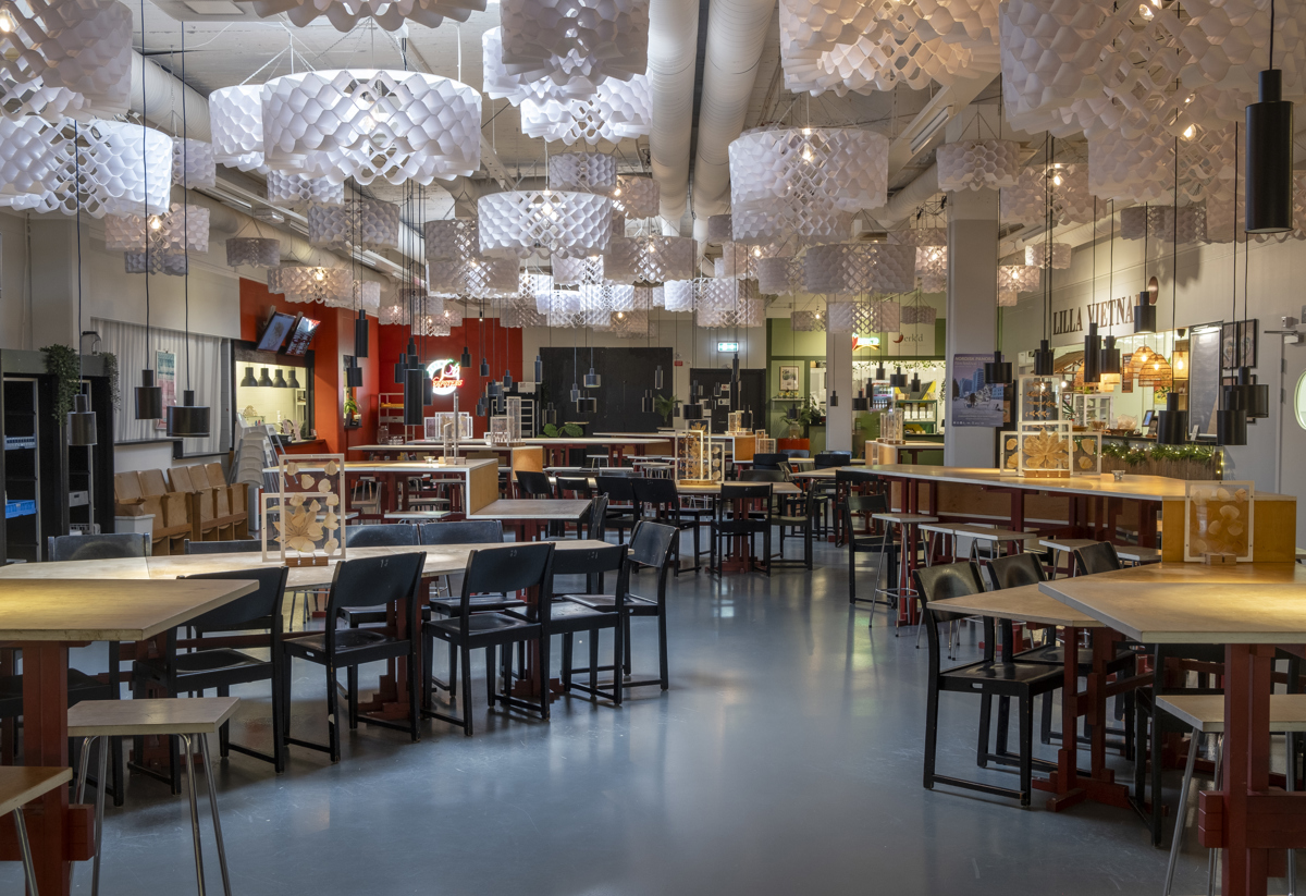 "Inomhusbild av en rymlig matsal med många bord och stolar. Taket är dekorerat med stora, vita lampor och lokalen är välbelyst med både naturligt och konstgjort ljus.