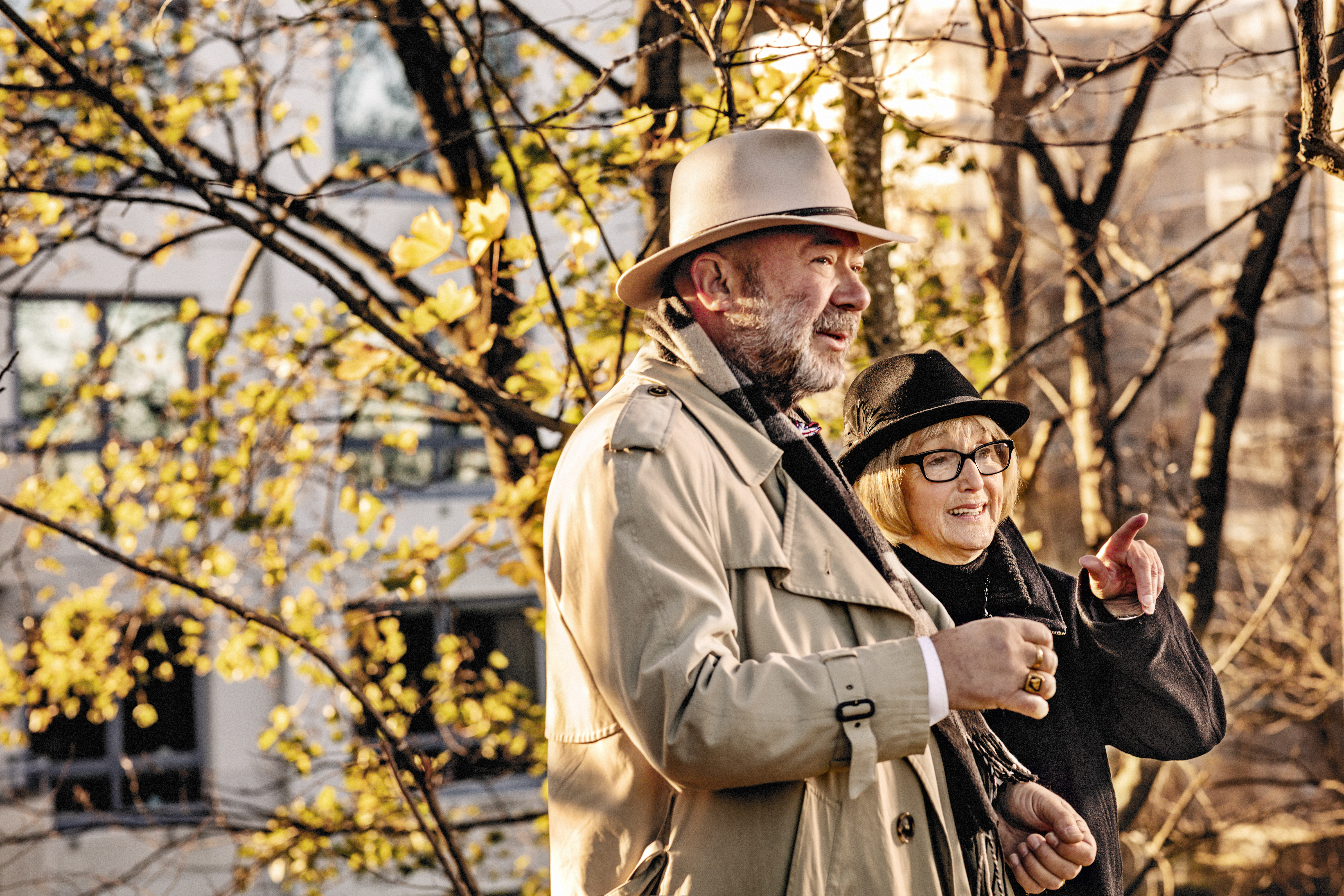 Äldre par, båda klädda i rock och hatt står framför ett höstigt träd, i bekgrunden ett flerfamiljsfastighet, 