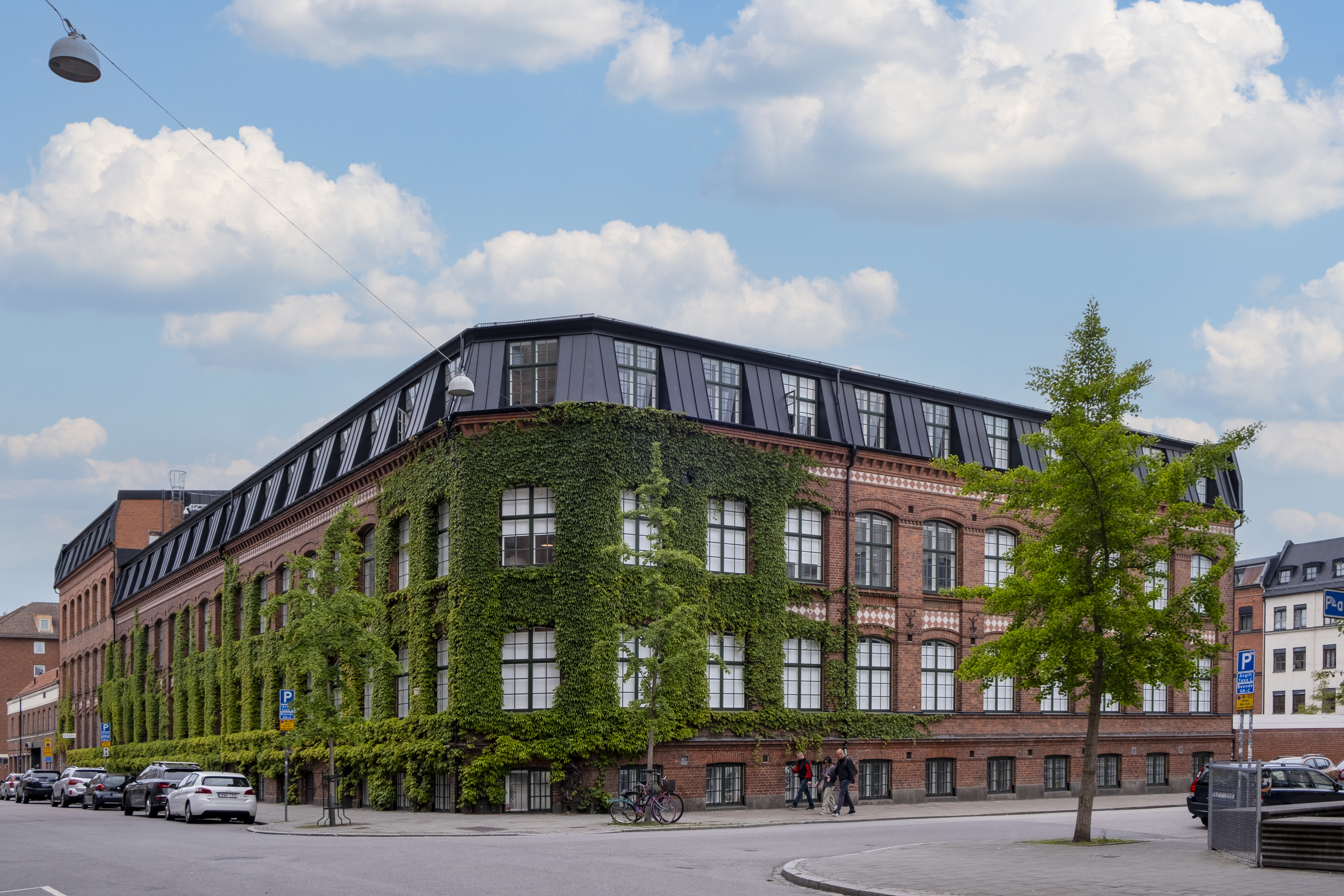 Ett stort tegelhus i hörnet av en stadsgata, med gröna klätterväxter som täcker delar av fasaden. Byggnaden har stora fönster och ett svart tak med moderna inslag. Framför huset finns några träd, parkerade bilar och människor som promenerar på trottoaren. Himlen är blå med vita moln.