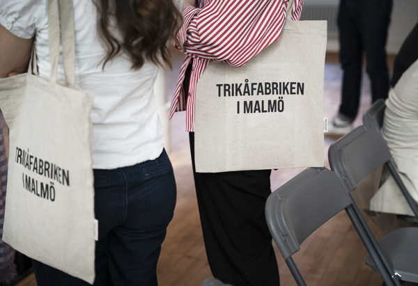 Två personer står bredvid varandra med tygkassar över axeln, båda med texten "TRIKÅFABRIKEN I MALMÖ" tryckt på väskan. De står i ett rum med hopfällbara stolar och trägolv.