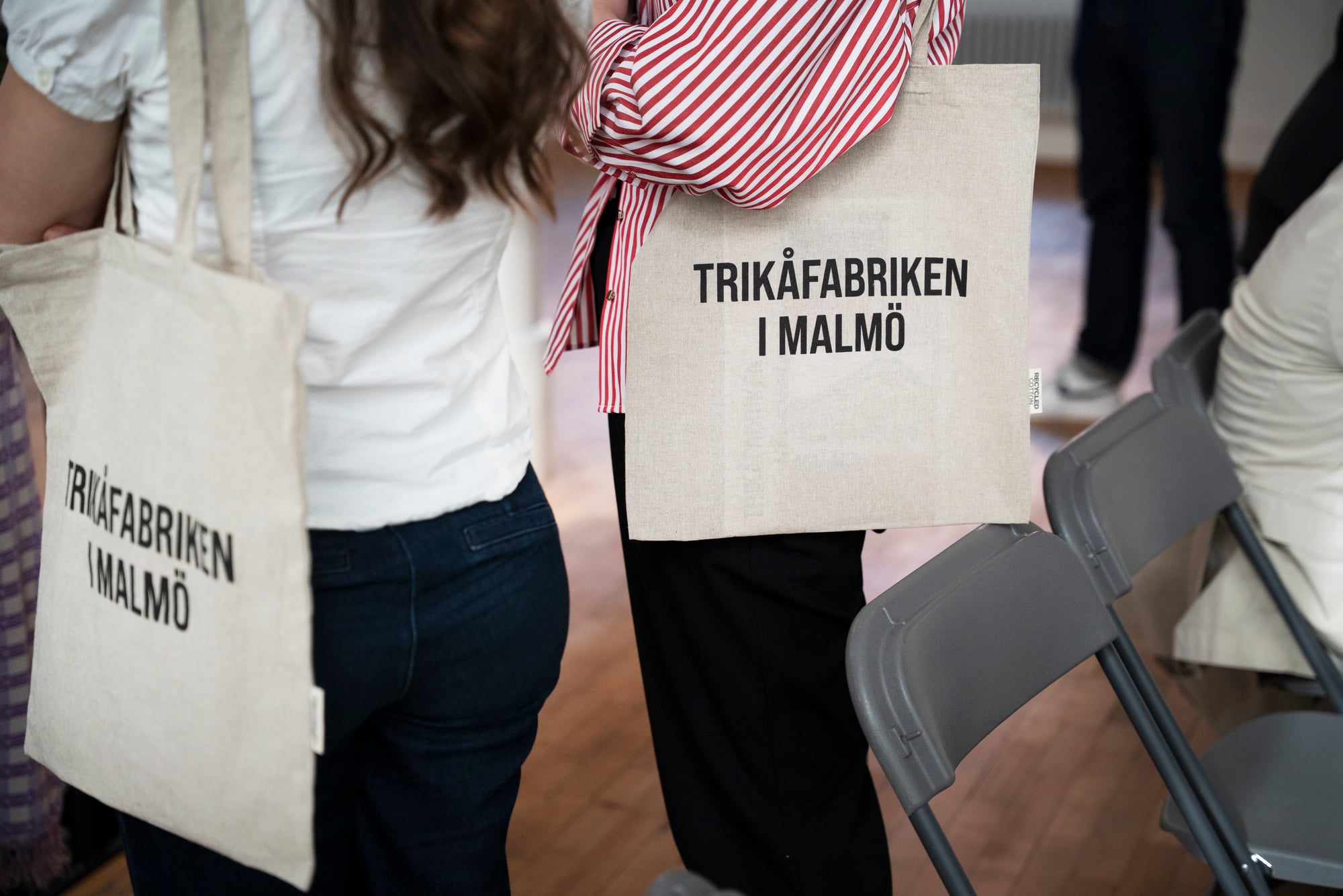 Två personer står bredvid varandra med tygkassar över axeln, båda med texten "TRIKÅFABRIKEN I MALMÖ" tryckt på väskan. De står i ett rum med hopfällbara stolar och trägolv.