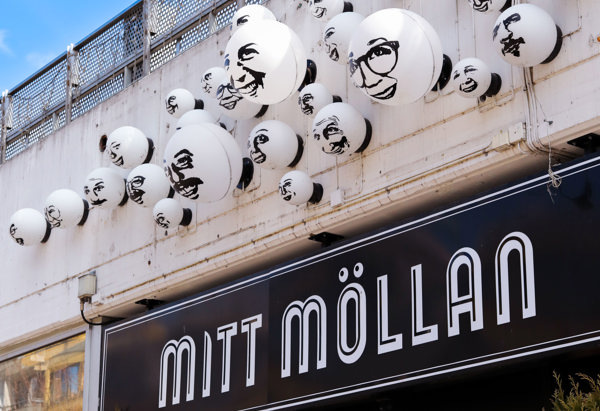 Exteriör av en galleria med en skylt som lyder 'MITT MÖLLAN'. Ovanför skylten finns många vita sfäriska objekt fästa på väggen, var och en med svarta illustrationer av ansikten.