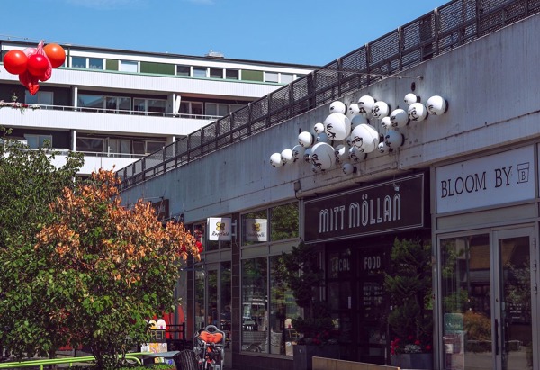 En utomhusscen i ett kommersiellt område med flera butiker. I förgrunden finns ett träbord med bänkar, och bakom det finns två en fasad till gallerian med skyltar som lyder 'MITT MÖLLAN' och 'BLOOM BY'. Ovanför butikerna finns en unik installation bestående av flera vita klot med illustrerade ansikten på. Till vänster flyger röda ballonger. I bakgrunden syns bostadshus med balkonger.