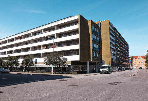 En modern flerfamiljsbyggnad med en gulbrun fasad och vita balkonger. Byggnaden är belägen vid ett gatuhörn med parkerade bilar och träd som kantar trottoaren. Himlen är klar och blå.