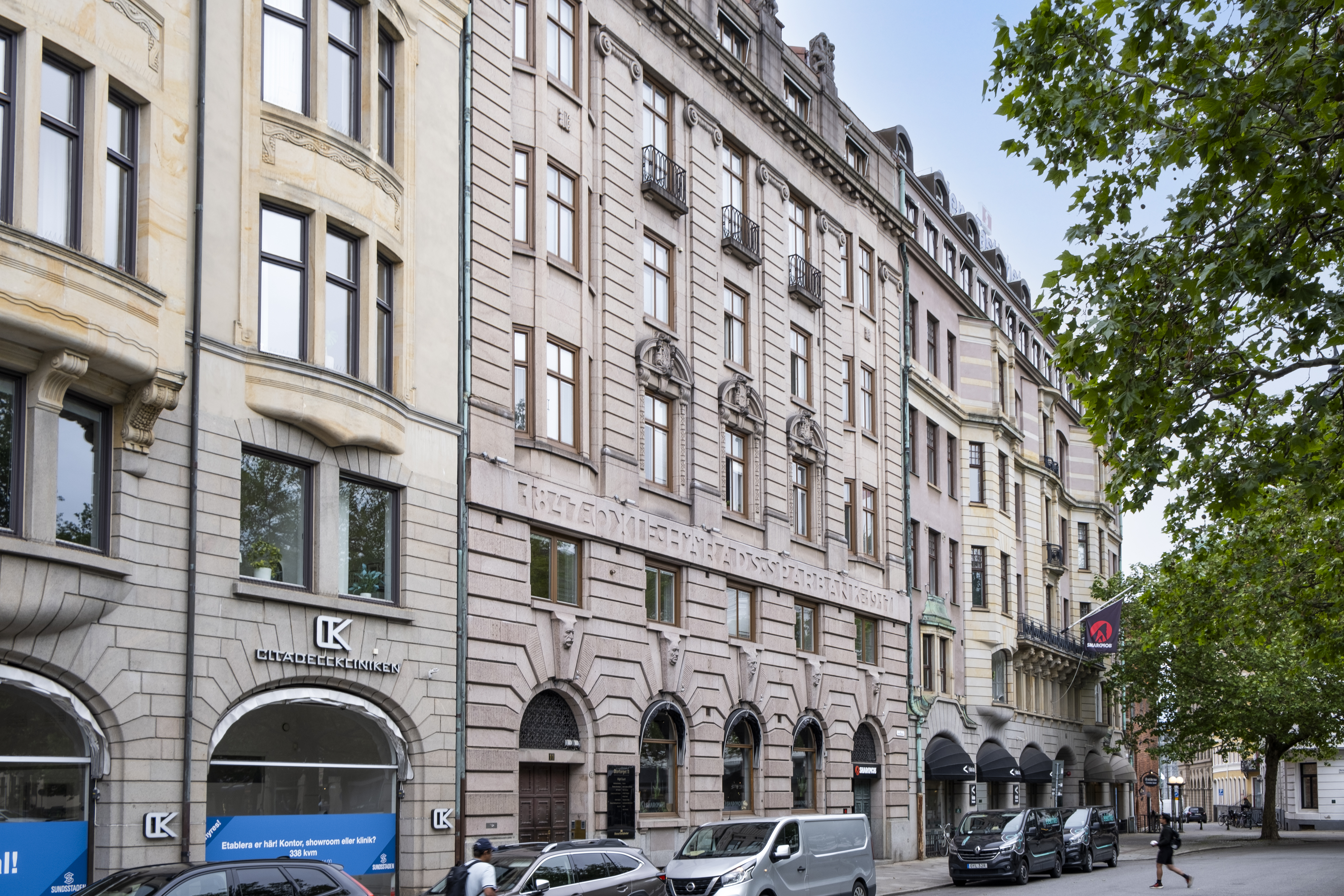 Fasad av en rad äldre stenbyggnader längs en stadsgata, med butiker i bottenplan, parkerade bilar vid trottoaren och gröna träd som ramar in gatan.