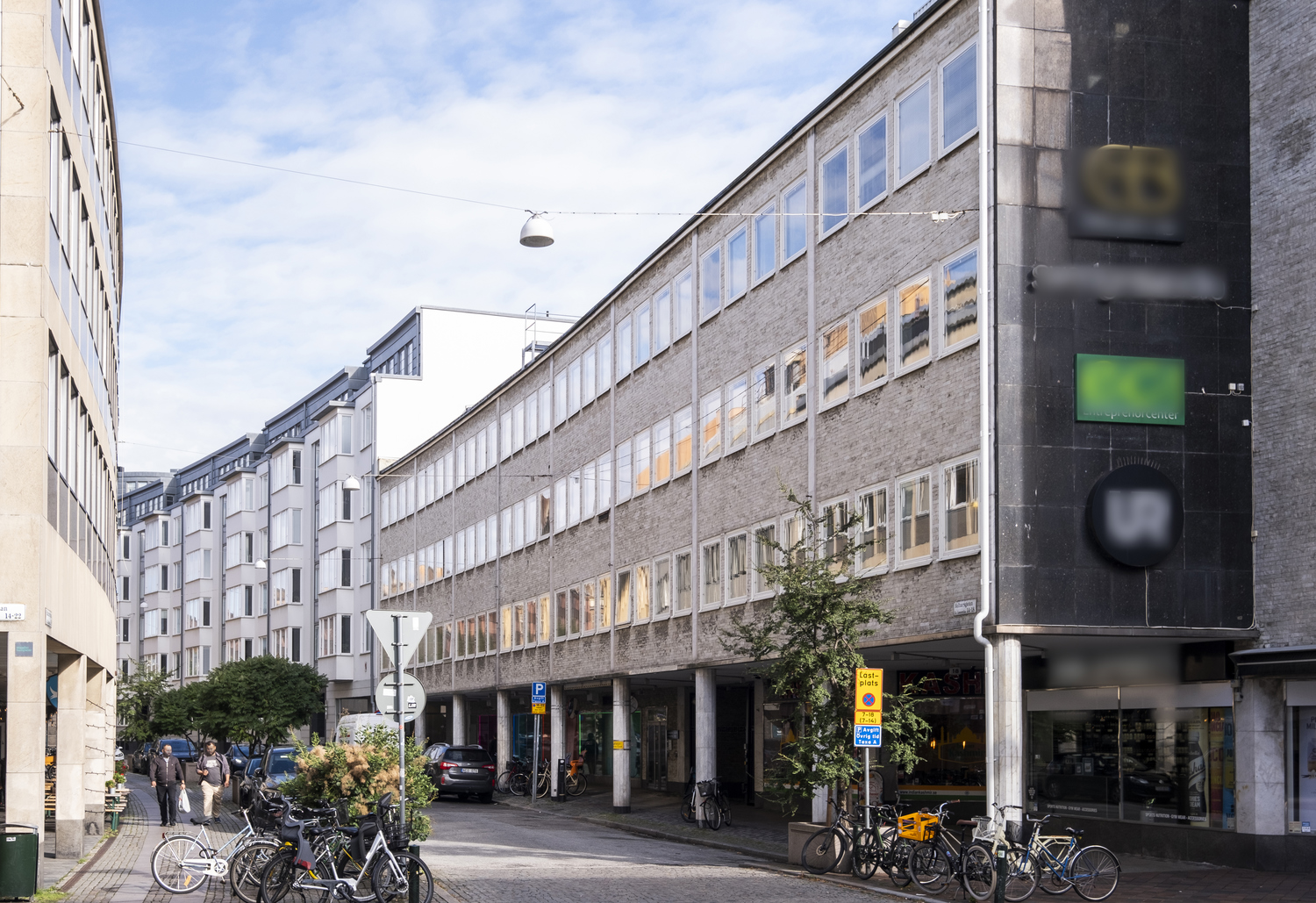 Fasadvy över ett äldre kontors- och butikshus i sten längs en smal stadsgata, med stora fönster i flera våningar, cyklar parkerade framför entréerna och människor som rör sig på trottoaren en solig dag.