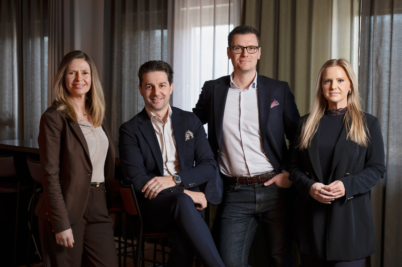 Fyra personer i affärskläder står tillsammans inomhus framför stora gardinklänlda fönster, och ser rakt in i kameran i en professionell gruppbild.
