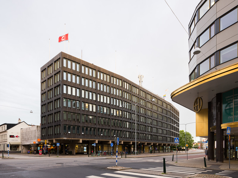 Fasadvy av ett större mörkbrunt kontorshus i flera våningar vid en gatukorsning, med pelare i bottenplan, många fönster och en flagga på taket, omgiven av trottoarer, skyltar och närliggande byggnader.