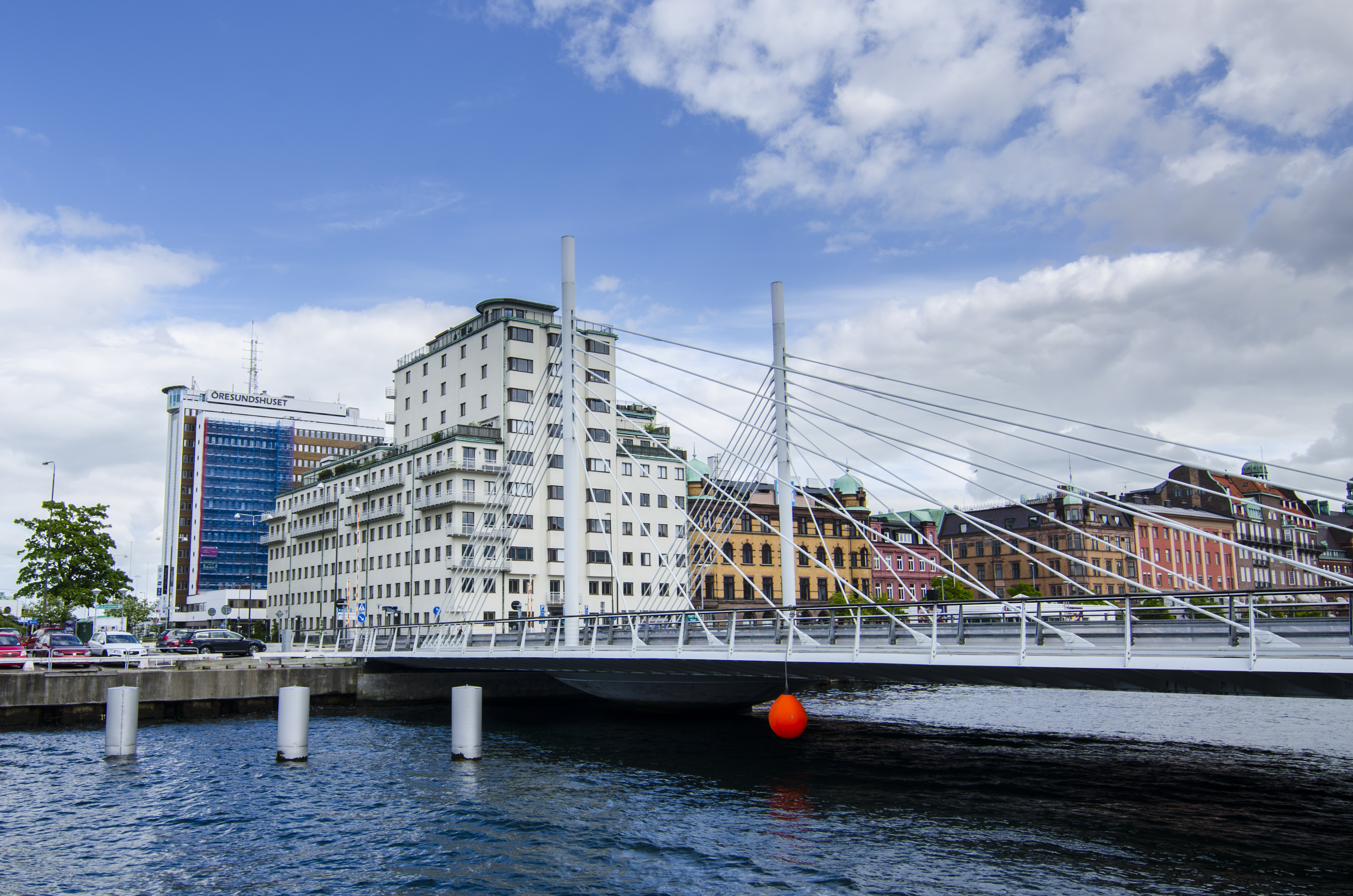 En modern hängbro över vatten med stadskvarter och färgglada äldre hus i bakgrunden under en molnig himmel.