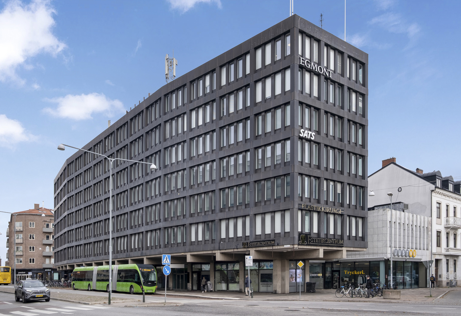 En mörkgrå kontors- och affärsbyggnad i ett gathörn med stora fönster, butiker i bottenplan och bussar samt cyklar längs den trafikerade gatan under en ljusblå himmel.