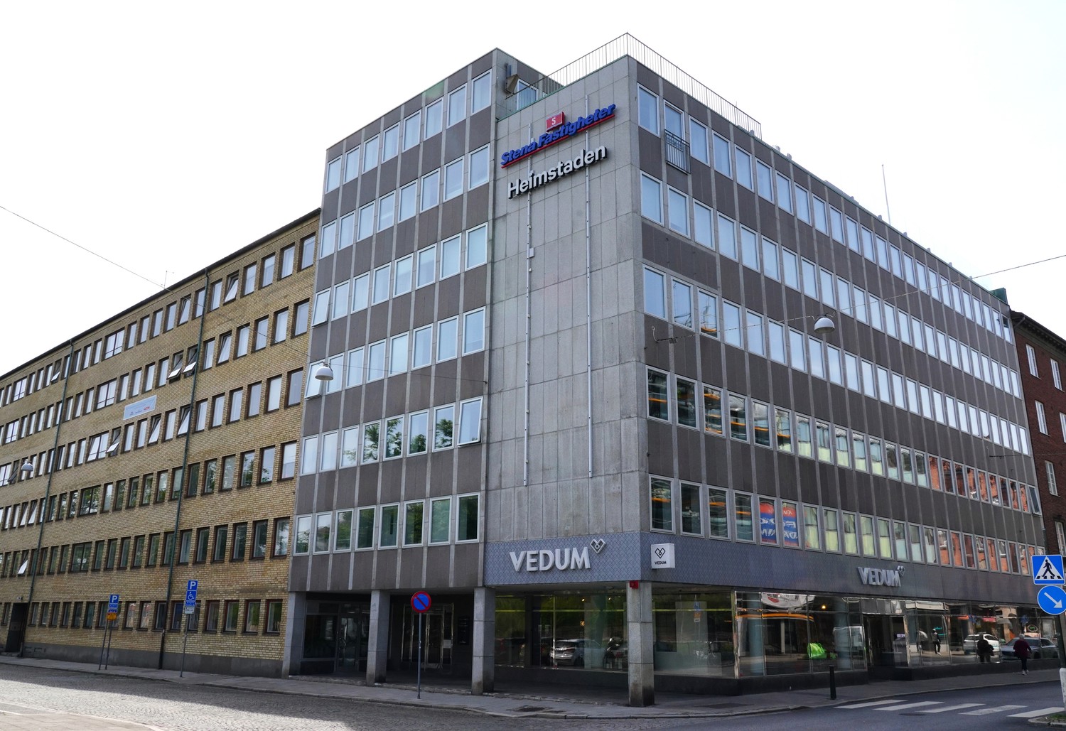 Fasadvy av ett större hörnhus i flera våningar med glas- och betongfasad, kontor ovanför och butiker i bottenplan, beläget vid en korsning med trottoarer, skyltar och tom gata i dagsljus.