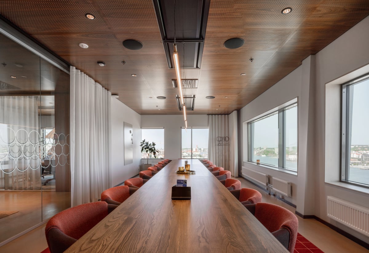 Ett elegant konferensrum med ett långt träbord omgivet av röda stolar. Stora fönster längs väggen släpper in naturligt ljus och erbjuder utsikt över staden. Taket är i trä med infälld belysning. Rummet har en modern och professionell atmosfär.