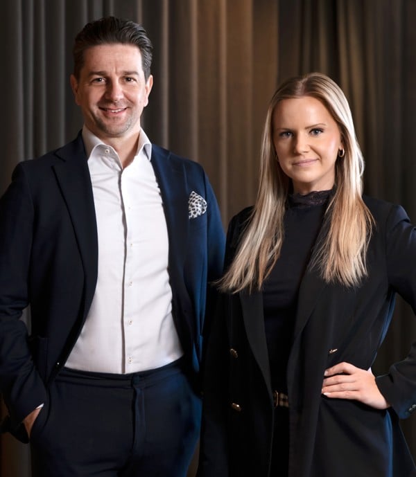 Två personer står bredvid varandra och tittar in i kameran. Personen till vänster bär mörk kostym och vit skjorta, och personen till höger bär svart kavaj och svart topp. Bakgrunden är mörk med gardiner.